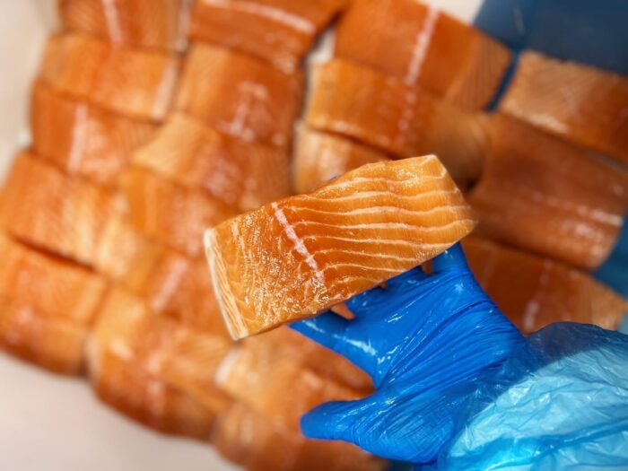 A hand in a blue protective glove holding a fresh salmon fillet above a blurred background of fresh salmon fillets